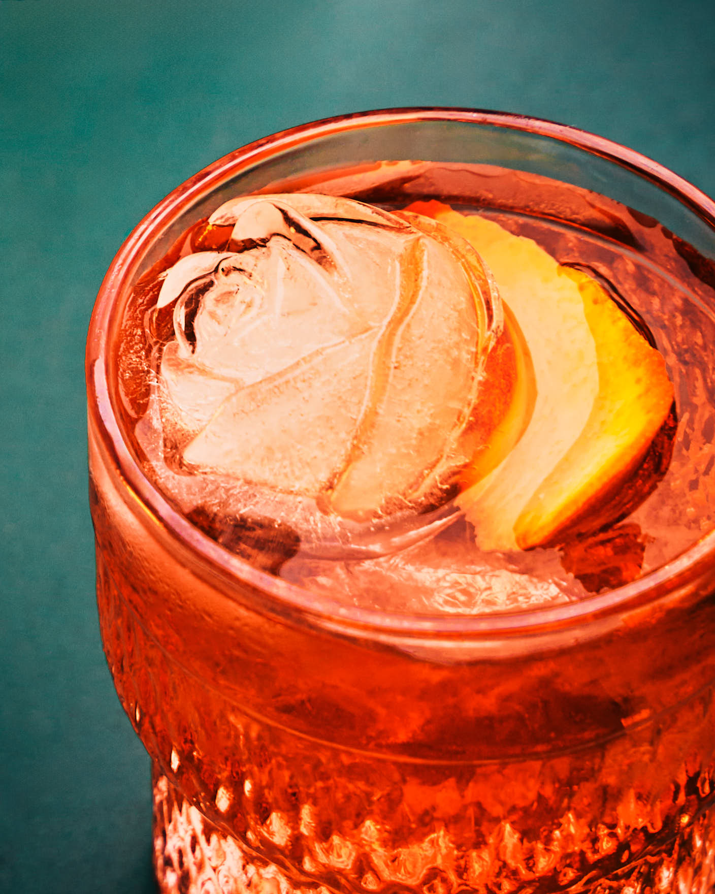 Close-up of an orange cocktail in a textured glass with a rose-shaped ice cube and a lemon peel garnish.
