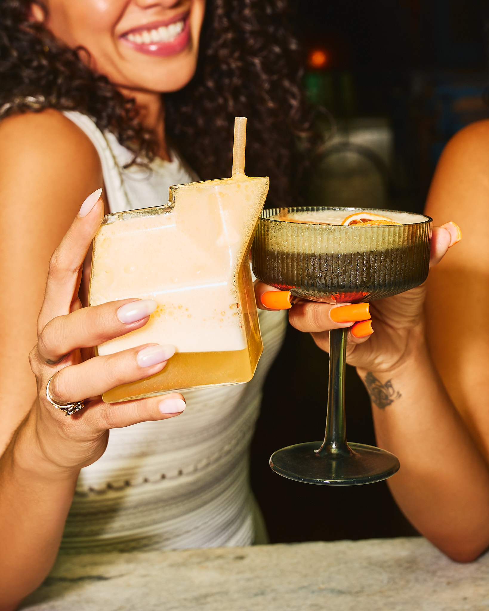 Two hands holding cocktails, one in a square glass with foam and a straw, the other in a stemmed glass with an orange garnish.
