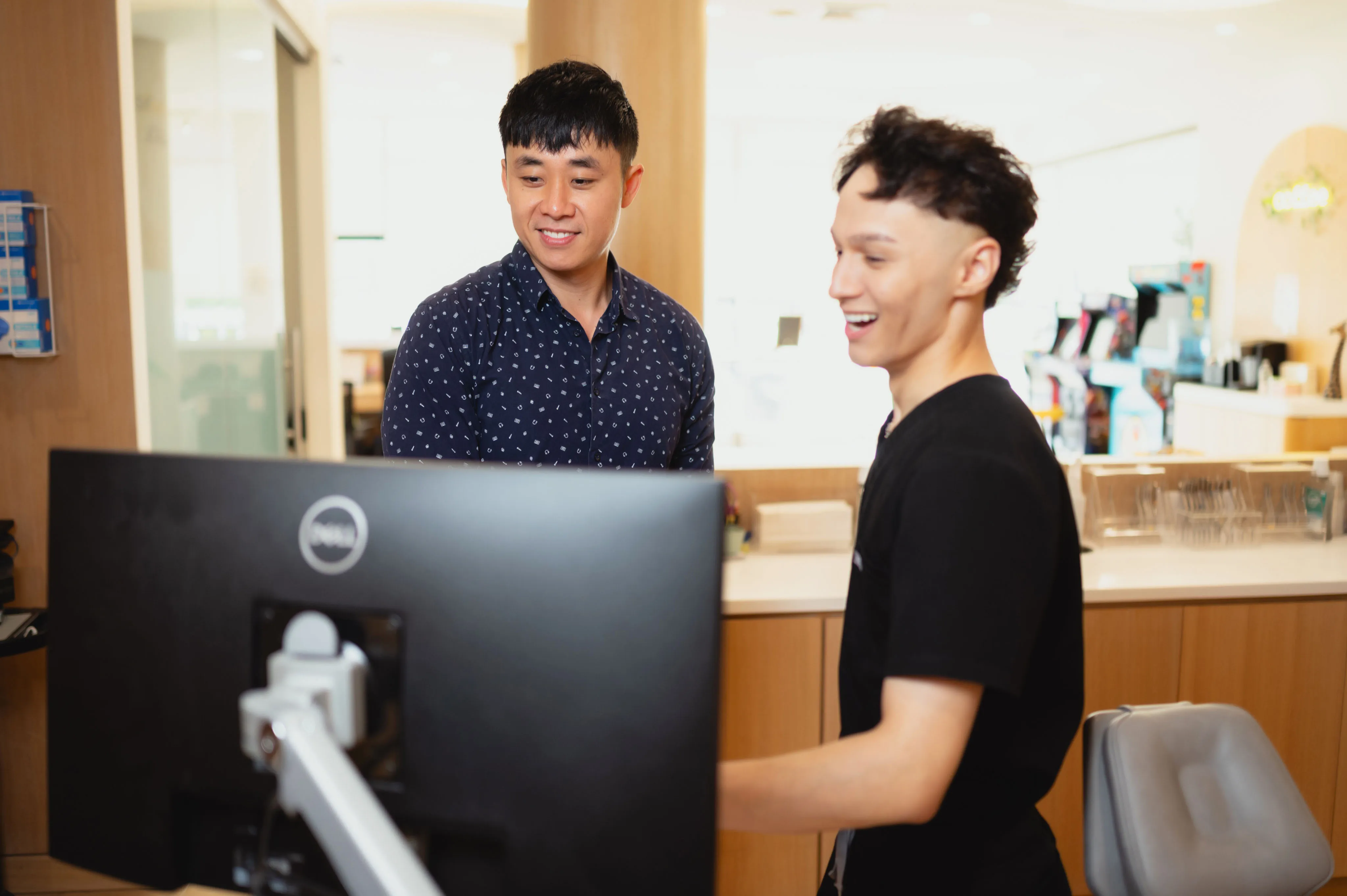 Two young men interacting in a dental office, one is standing and looking at the other who is seated at a desk with a computer. The atmosphere appears friendly and engaging.