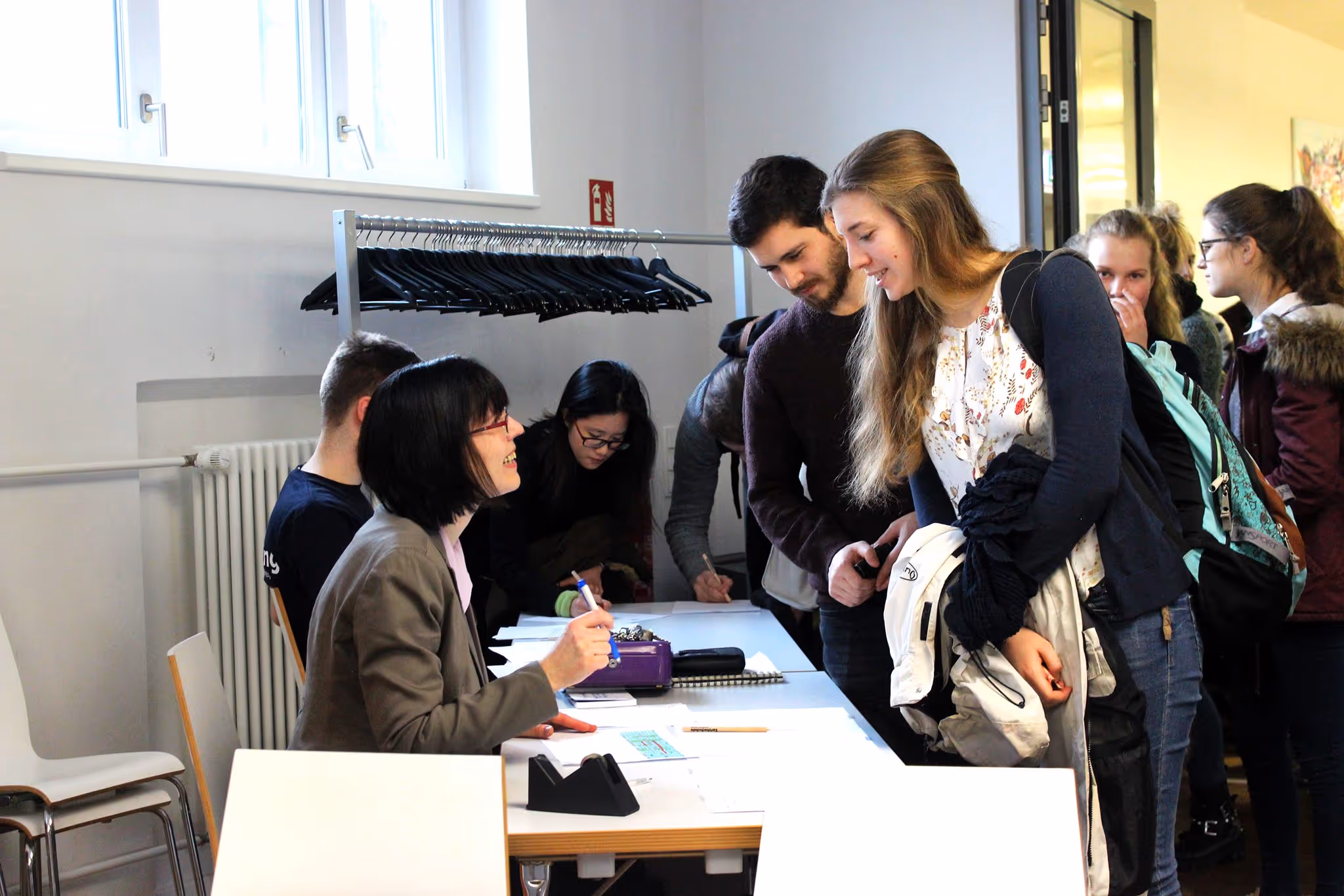 A group of business professionals or students collaborating around a table in an office setting, with one person pointing at a document.