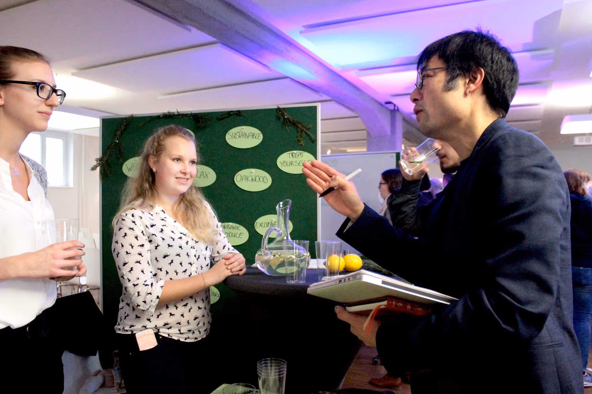 Two students or colleagues are talking and collaborating around a table in a brightly lit room with decorative green elements on the wall.