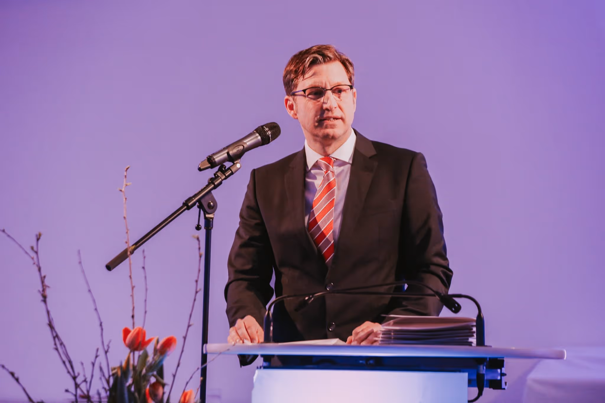 A young man in a suit is speaking at a lectern with a microphone, presenting to an audience in a formal setting.