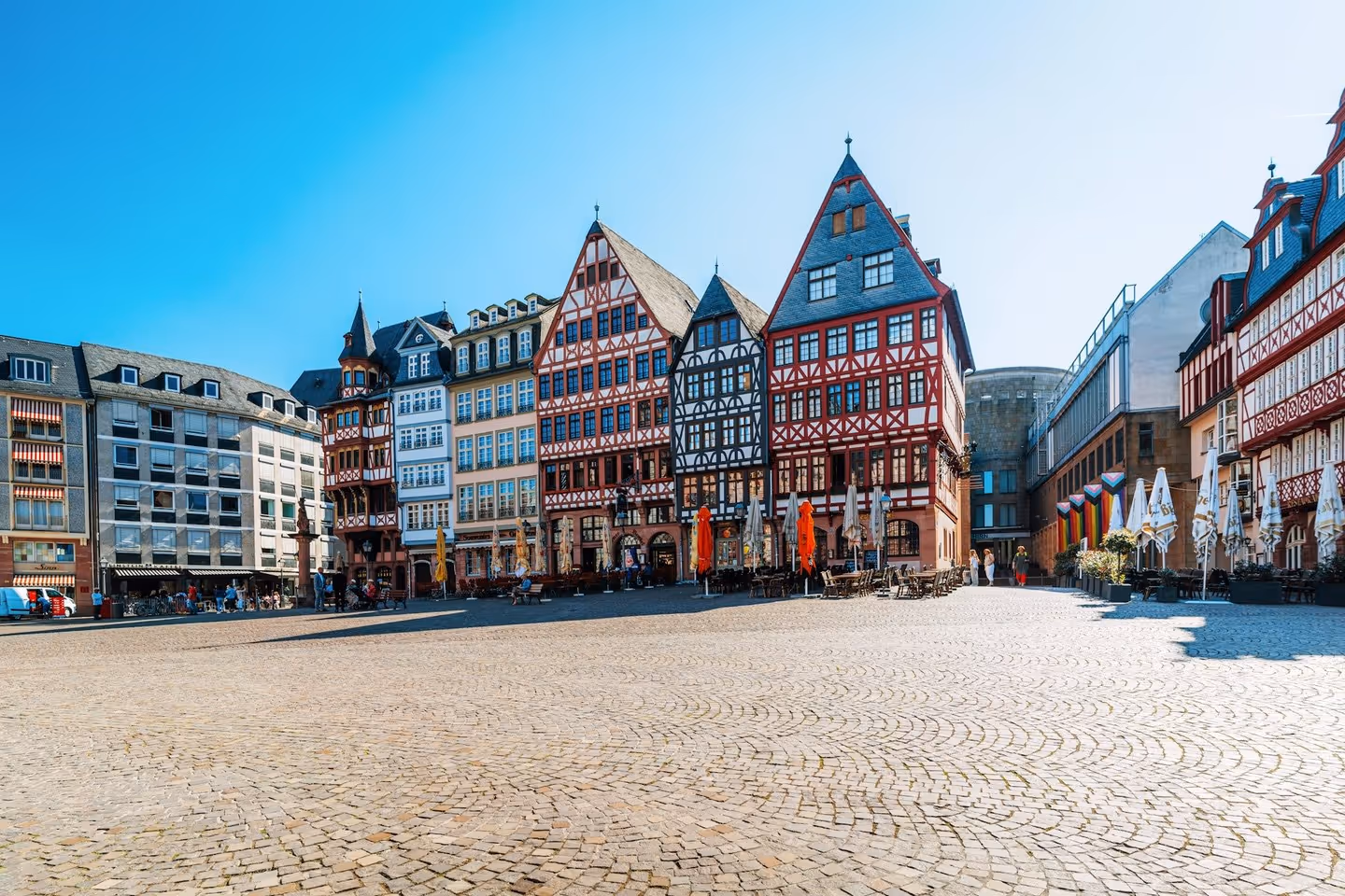 A daytime wide shot of a European square, featuring brightly colored half-timbered houses and a fountain in the foreground.