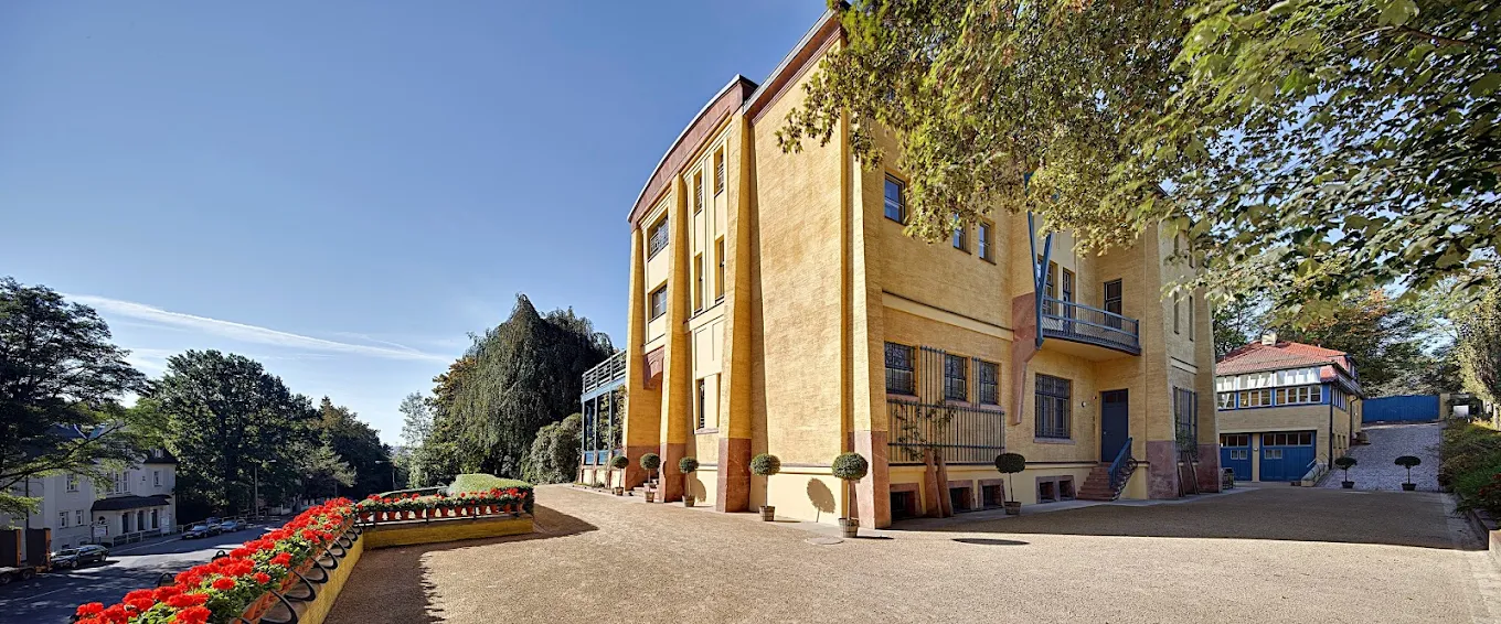 A historic, stately villa or large house at the end of a long driveway, surrounded by trees and illuminated by outdoor lights at dusk.