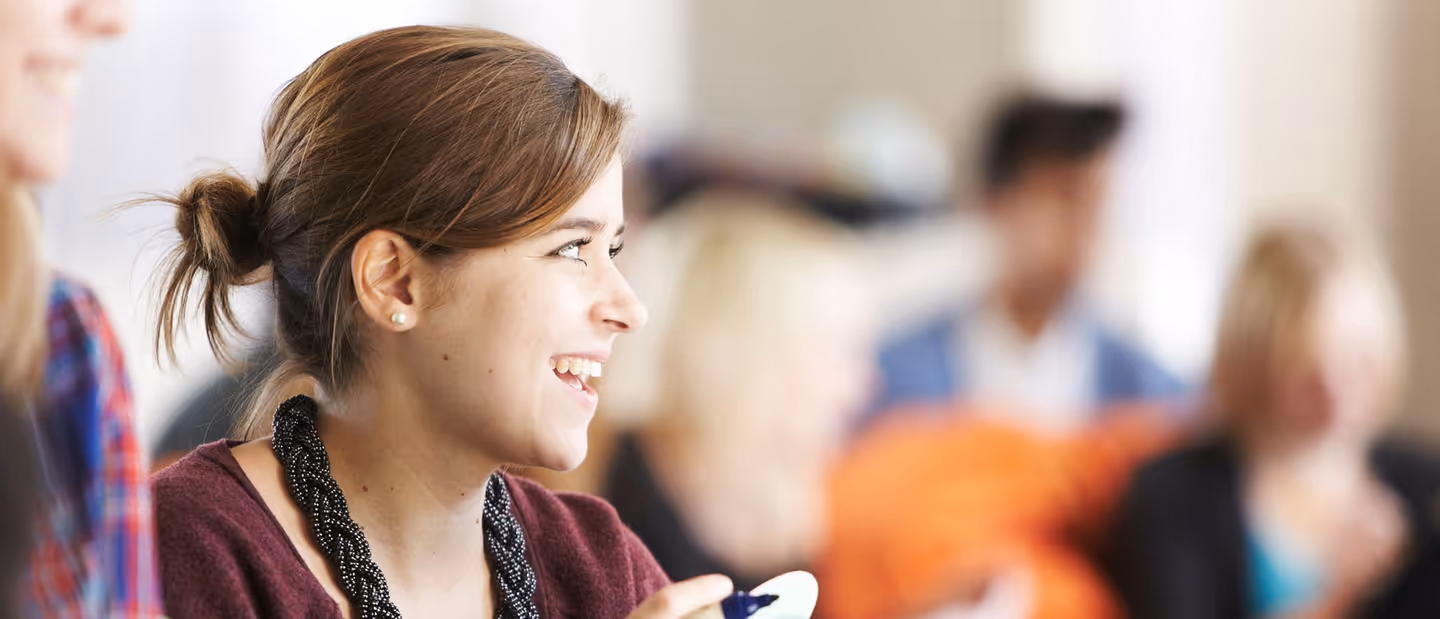 A close-up, candid shot of a young female student with long hair smiling at someone off-camera in a crowded setting.