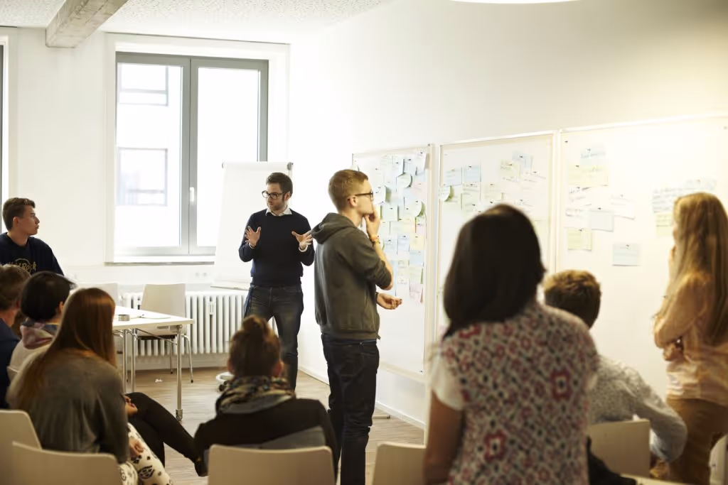 A small group of people seated in a relaxed, semicircle arrangement during an orientation or workshop, with a speaker in the background.