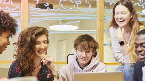 A bright, candid shot of two young female students smiling and working together on laptops in a modern classroom or study space.