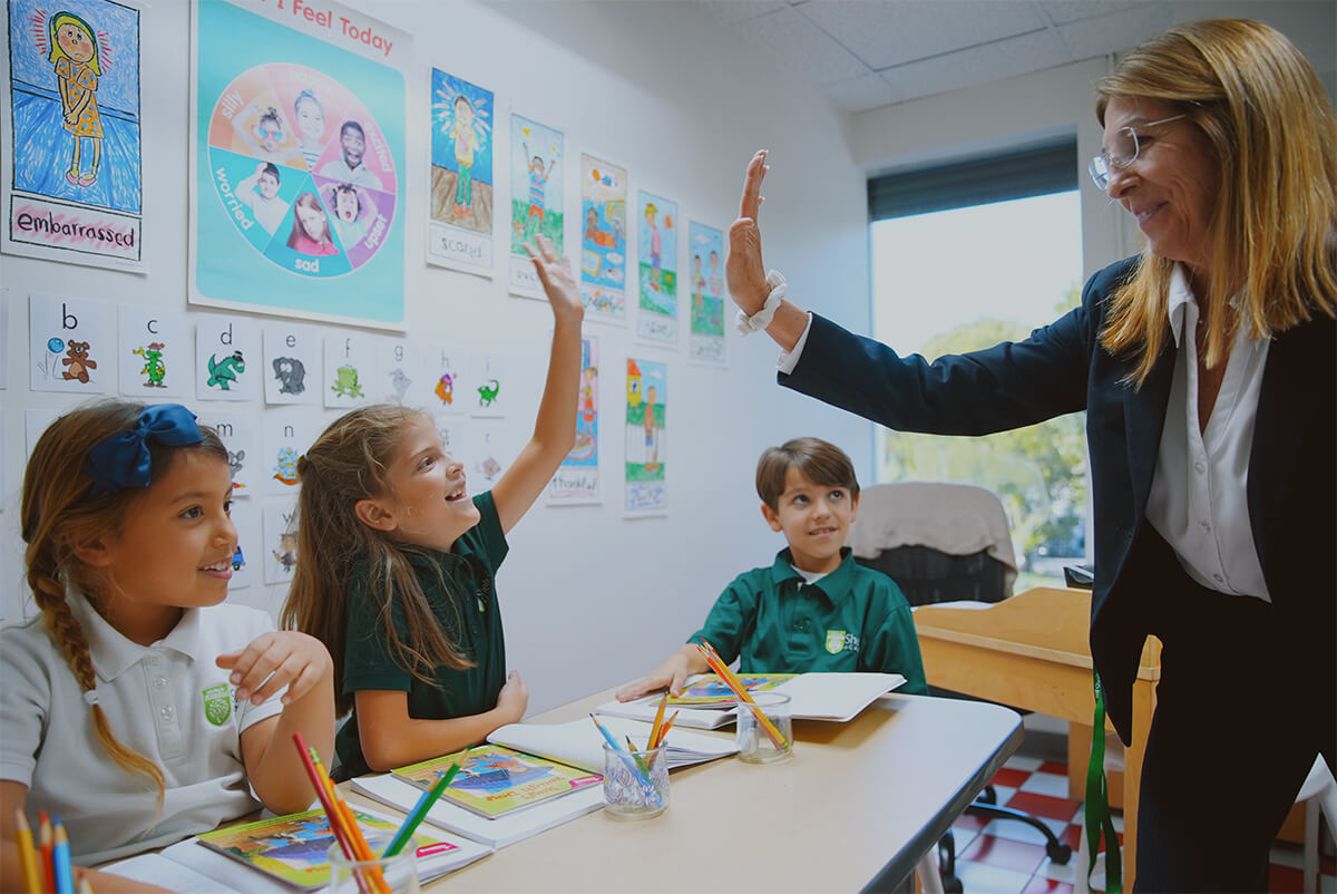 Teacher giving a high five to a smiling student in a classroom with three children sitting at a desk with books and pencils.