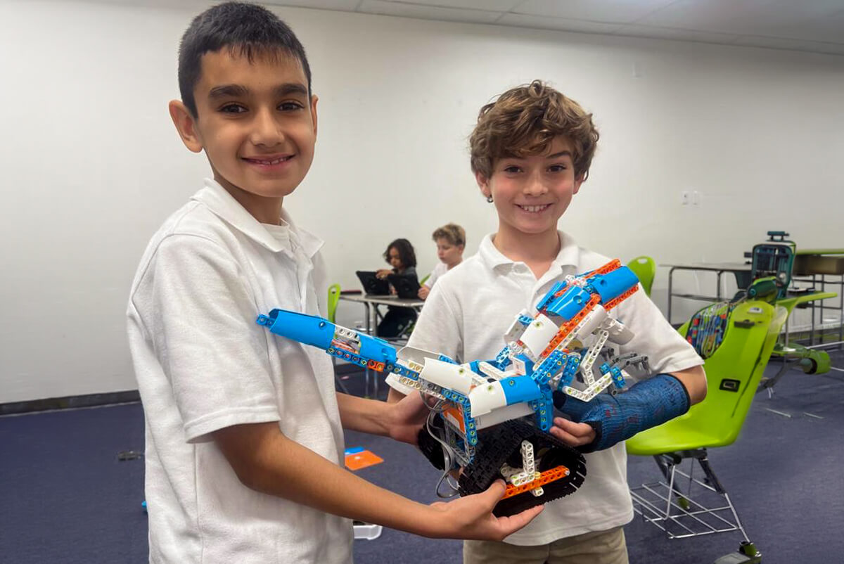 Two smiling boys holding a blue and white LEGO robotics model in a classroom.
