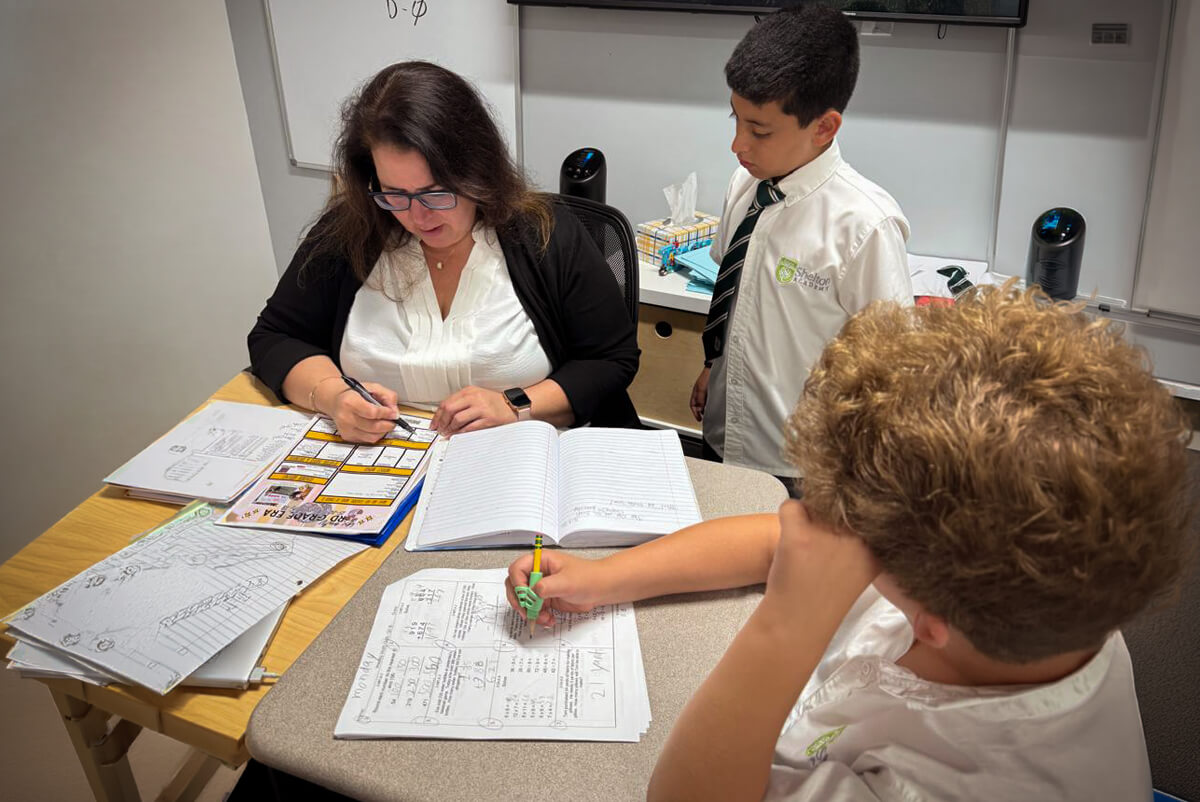 Teacher sitting at a desk with papers and folders, writing on a worksheet while a boy stands beside her and another boy with curly hair works on a worksheet at the table.