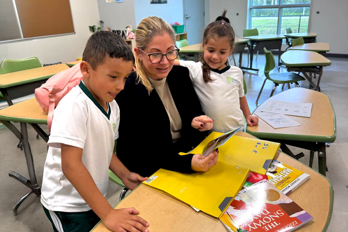 Teacher sitting at a classroom desk reading a book with two young students standing beside her.