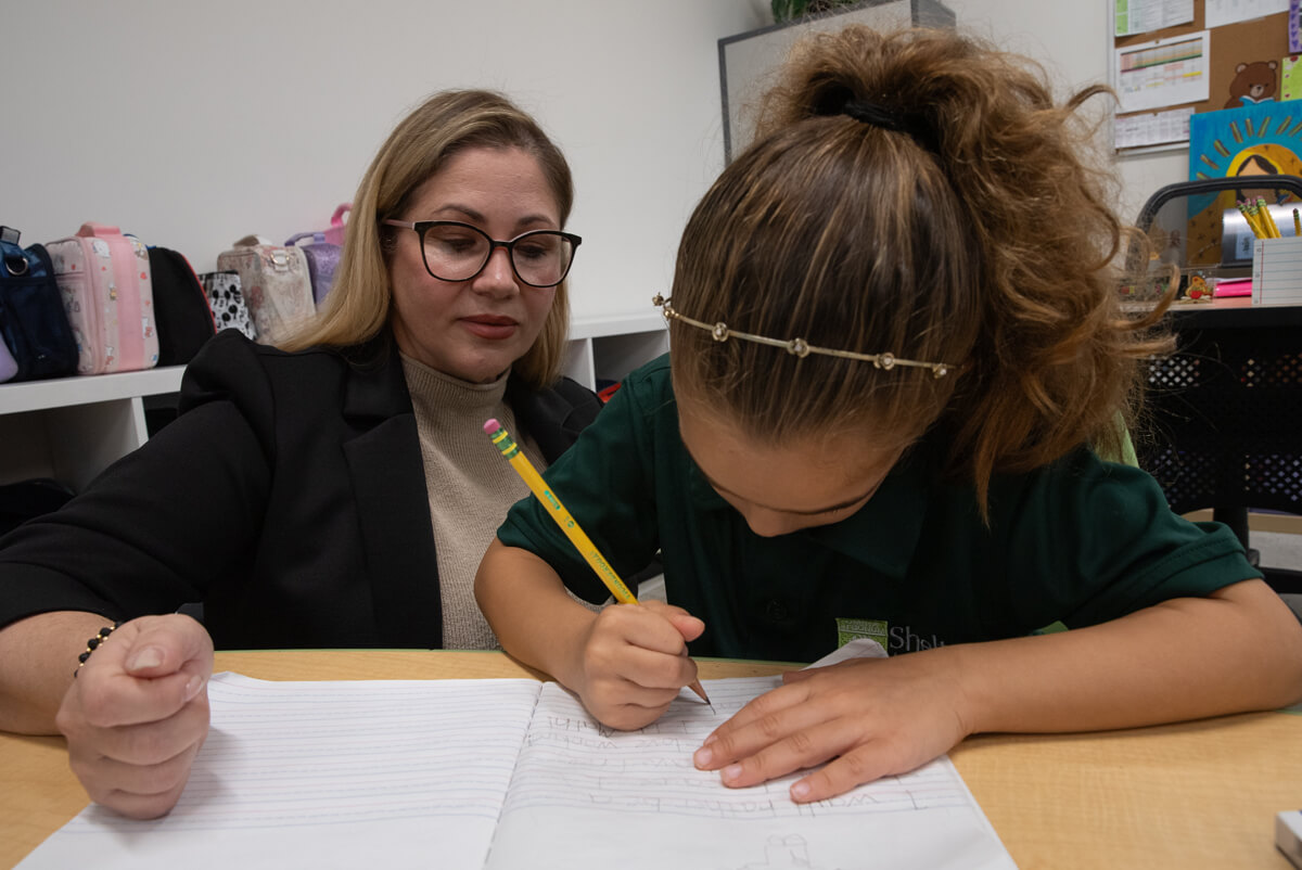 Teacher with glasses watching a young girl with a headband writing with a pencil in a notebook at a desk.