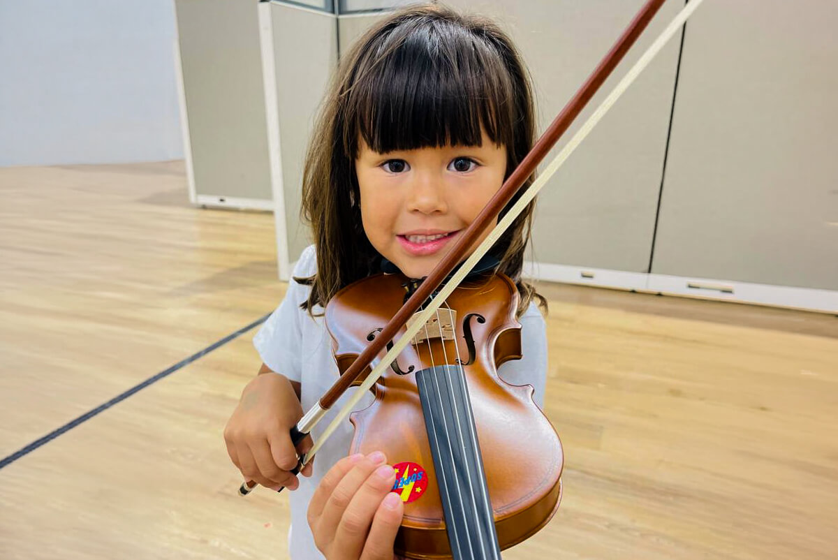 Young girl with dark hair playing a violin indoors on a wooden floor.