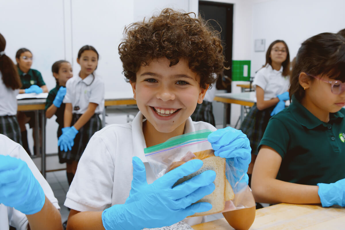 Smiling boy wearing blue gloves holding a sandwich in a plastic bag at a classroom table with other children.