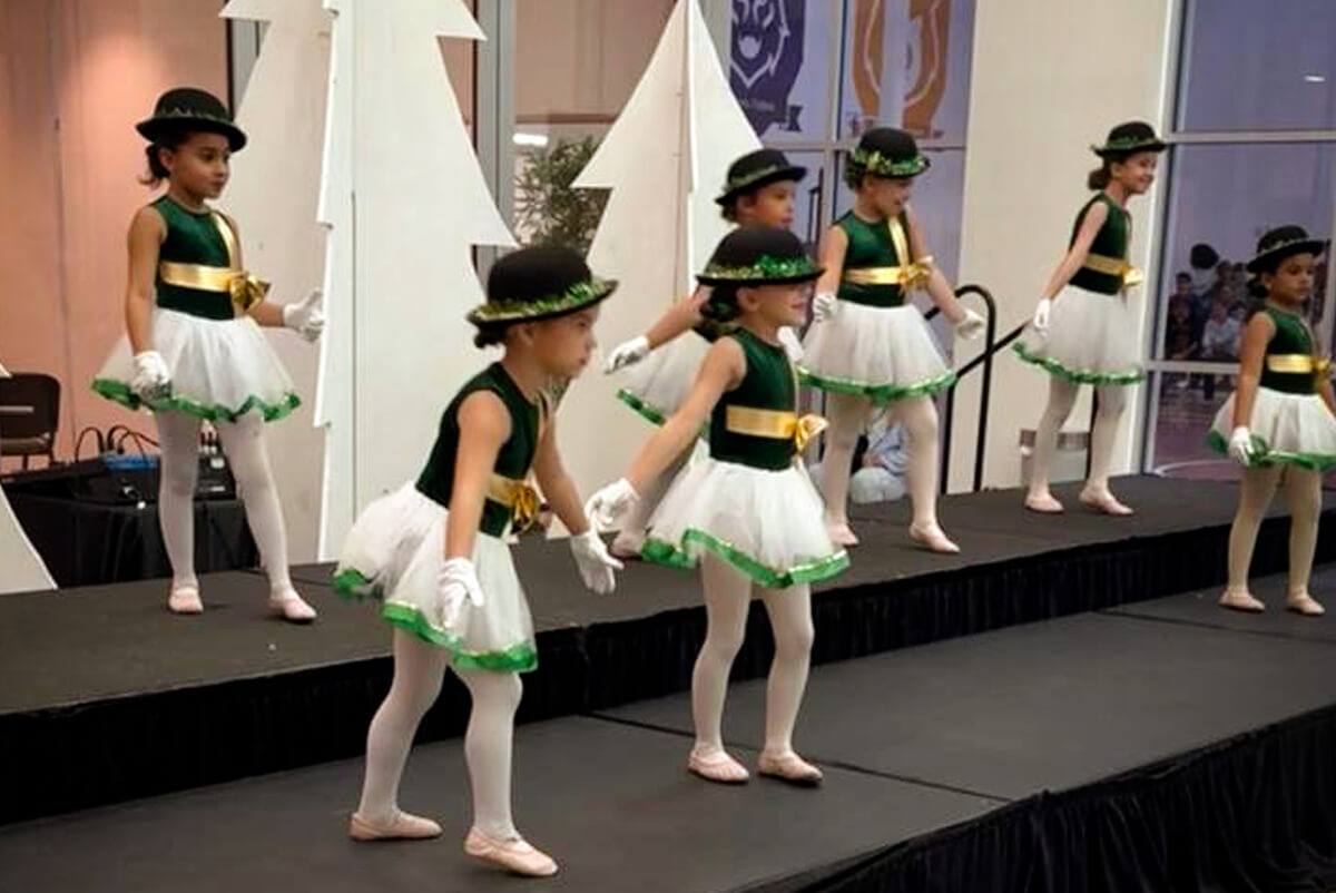 Young girls in green and white ballet costumes with black hats performing on a stage.