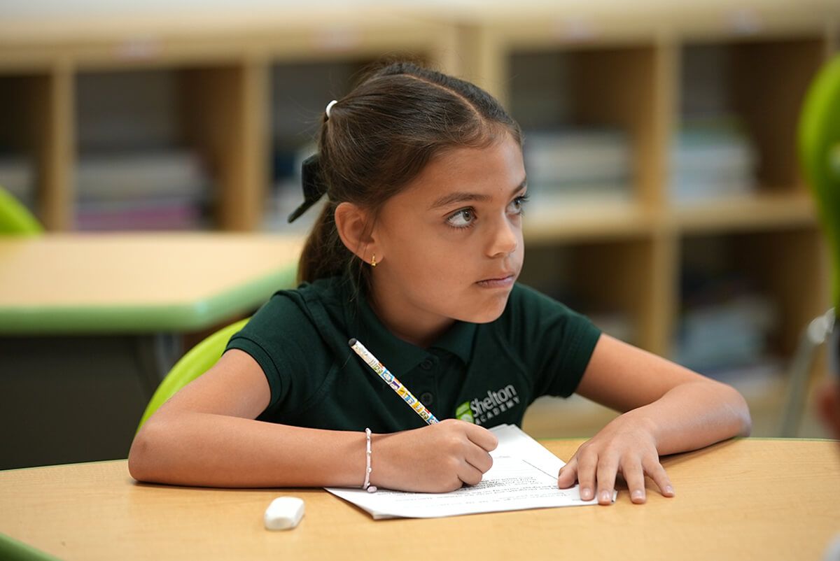 Young girl in green Shelton Academy polo shirt writing on paper at a classroom desk.