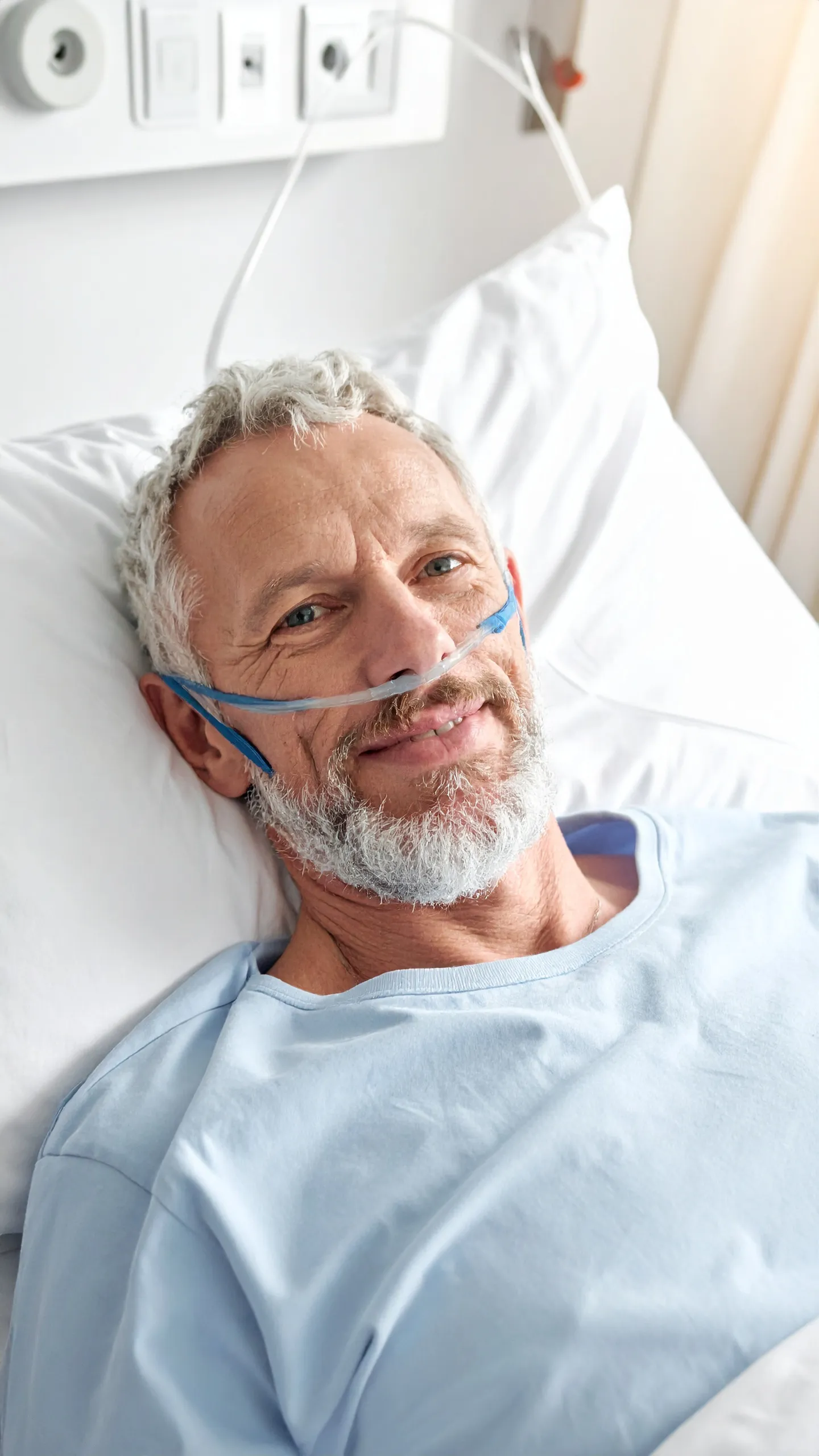 Older man with gray hair and beard lying in hospital bed wearing nasal oxygen tube and light blue hospital gown.