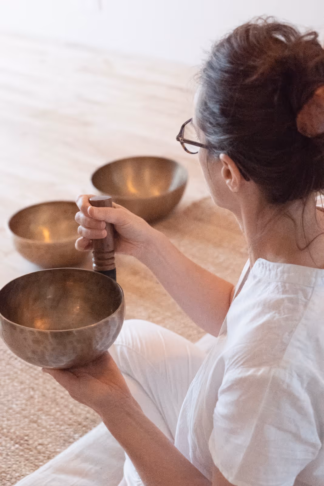 Femme en chemise blanche jouant d'un bol chantant en métal avec un maillet en bois, assise sur un tapis.