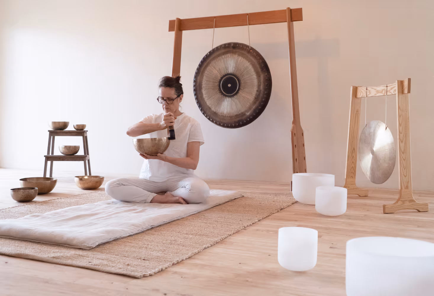 Femme assise en tailleur dans une pièce minimaliste jouant d’un bol chantant, entourée de bols chantants, gongs et instruments de méditation.
