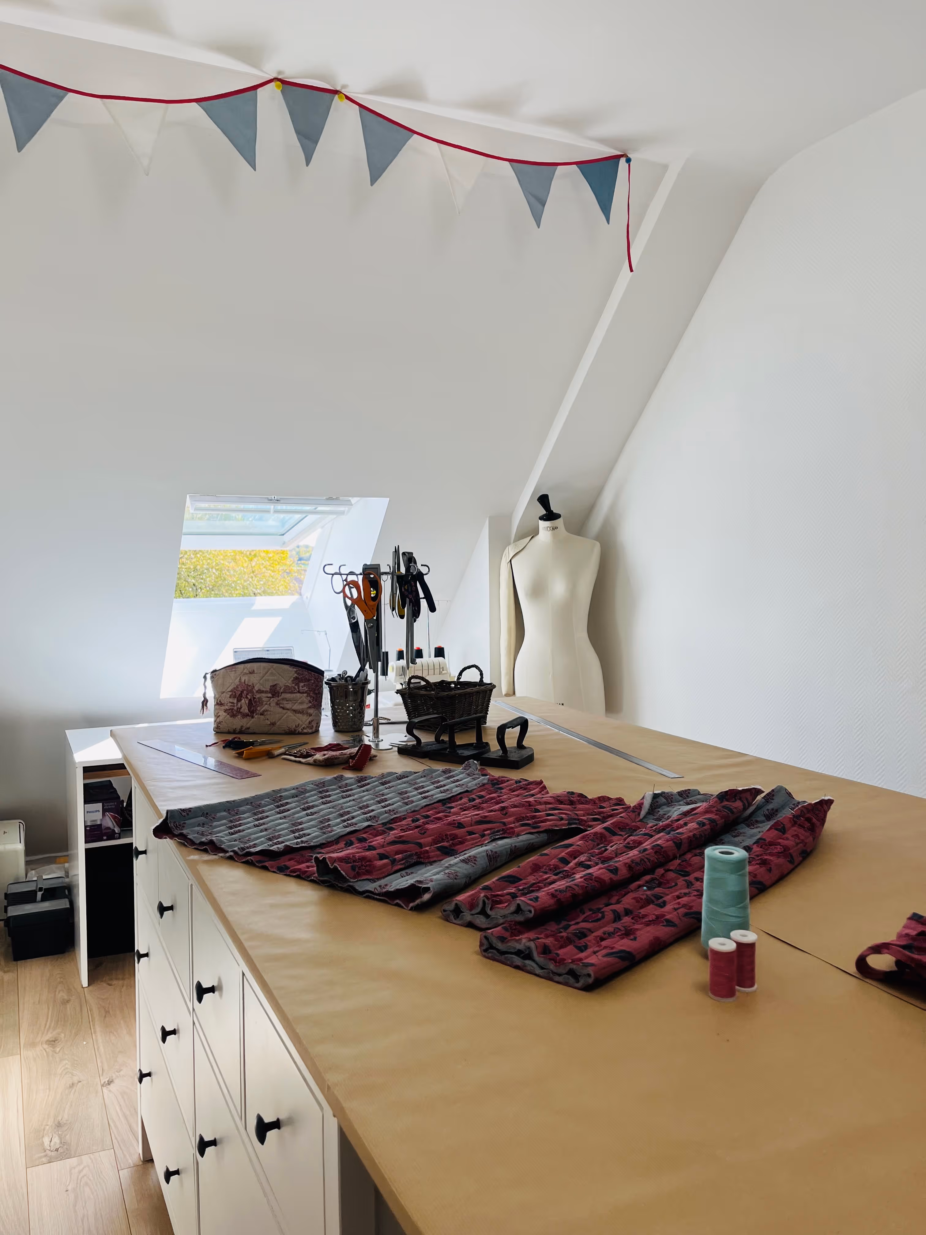 Table de couture avec tissus, outils, mannequin et fenêtre sous un plafond en pente.