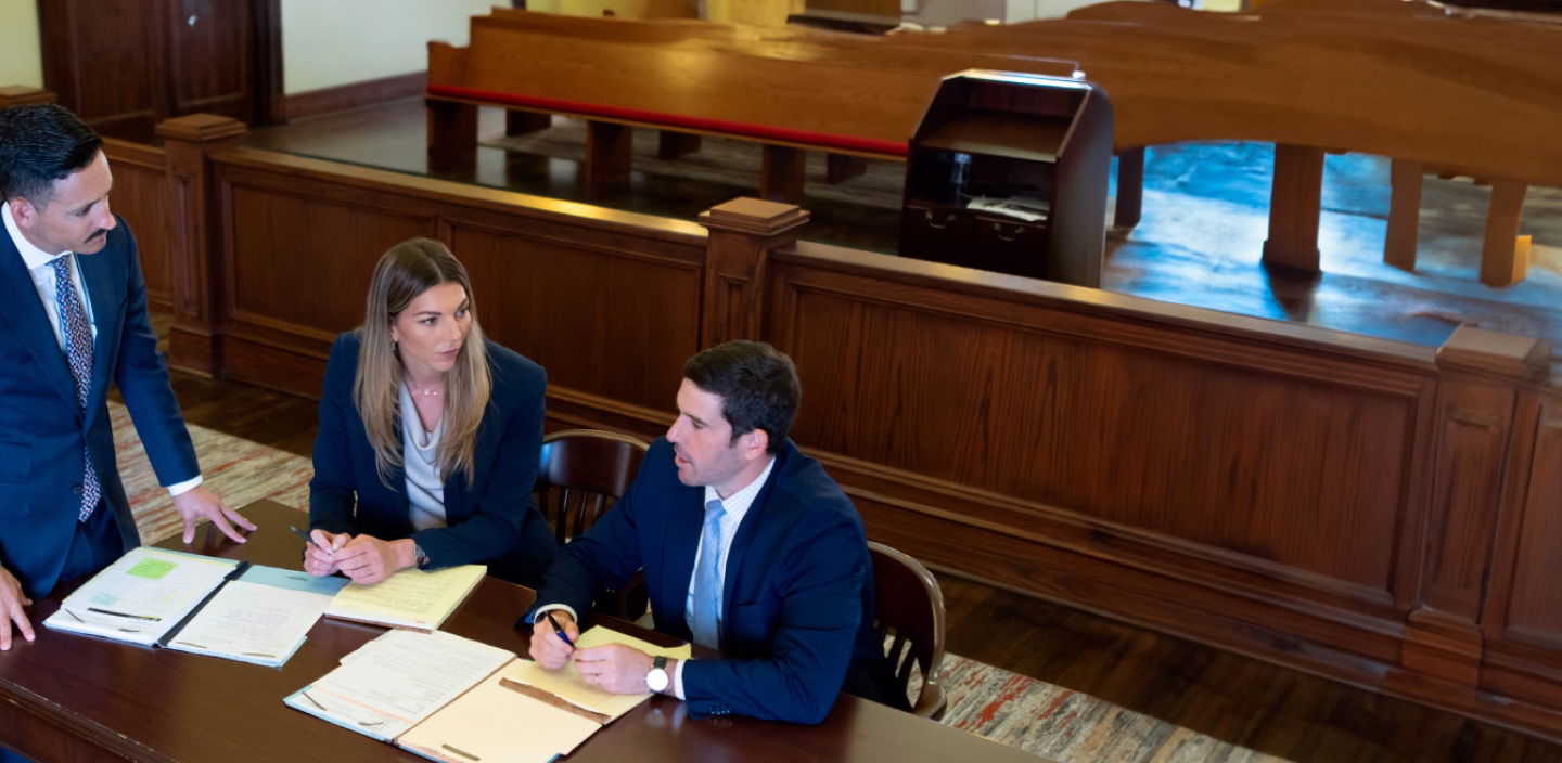 Three people in a courtroom discussing documents at a wooden table.
