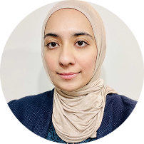 Portrait of a woman wearing a beige hijab and navy blue top, smiling softly against a light background.