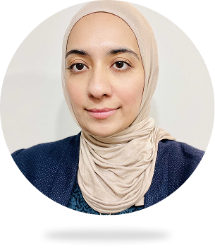 Portrait of a woman wearing a beige hijab and navy blue sweater against a plain light background.