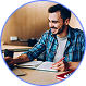 Man smiling while writing or working at a wooden desk with a laptop and papers.