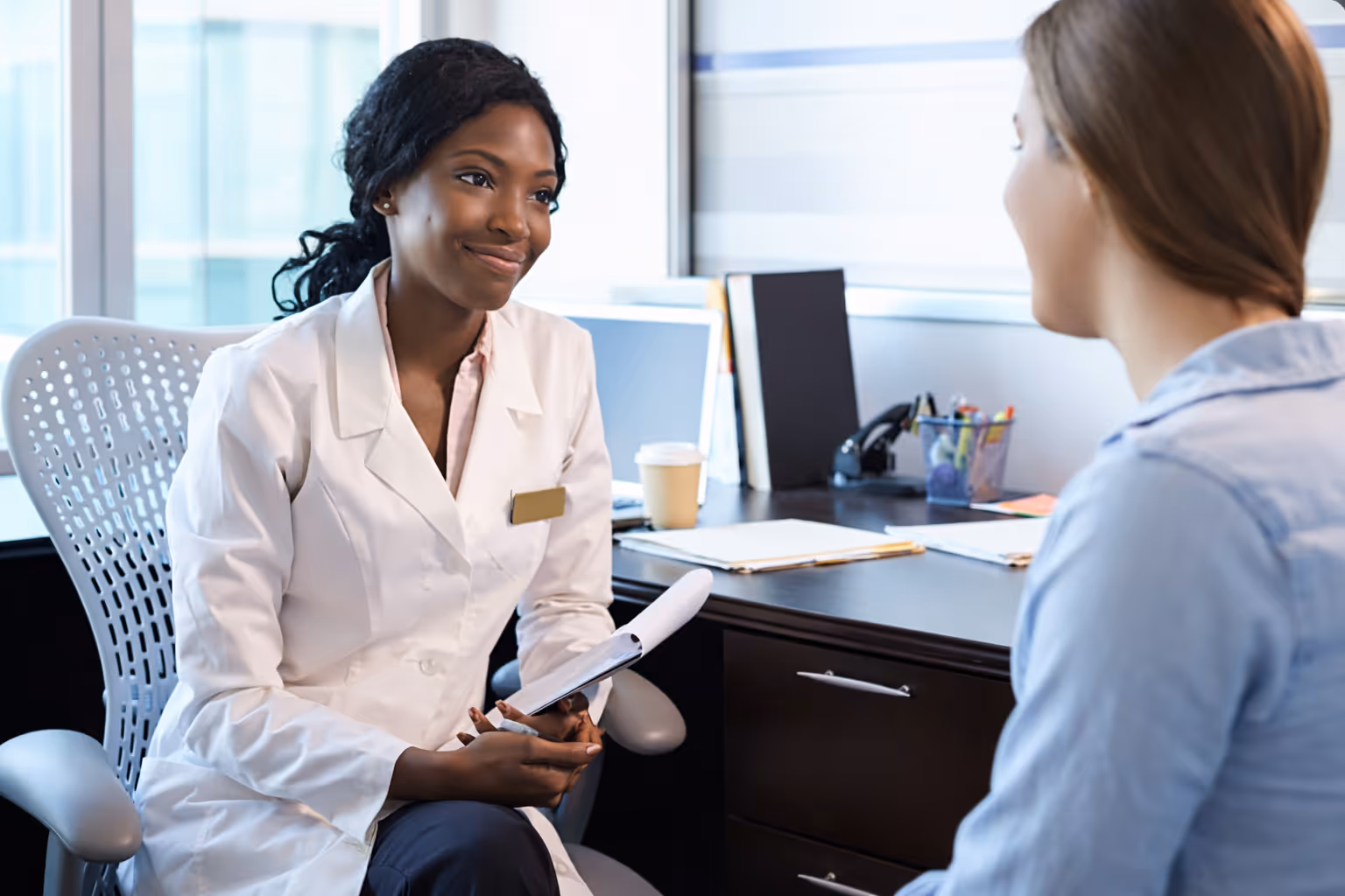 Female doctor in white coat sitting across from a female patient during a consultation, smiling