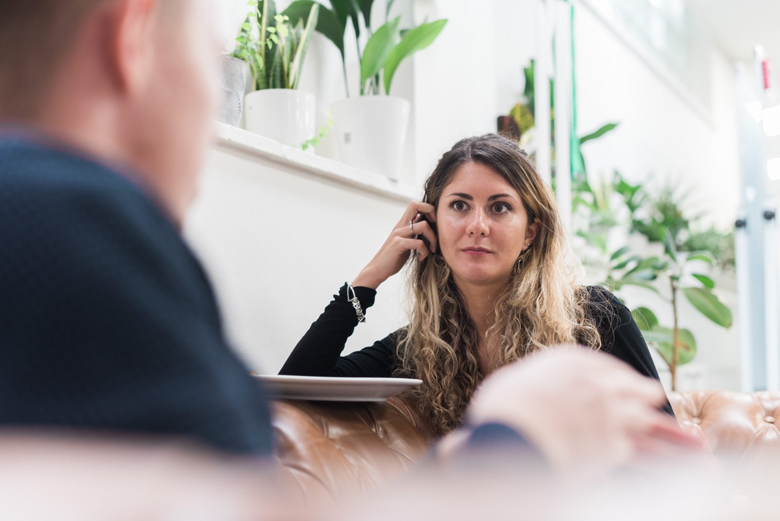 A woman with long hair listens intently during a conversation in a room with plants.