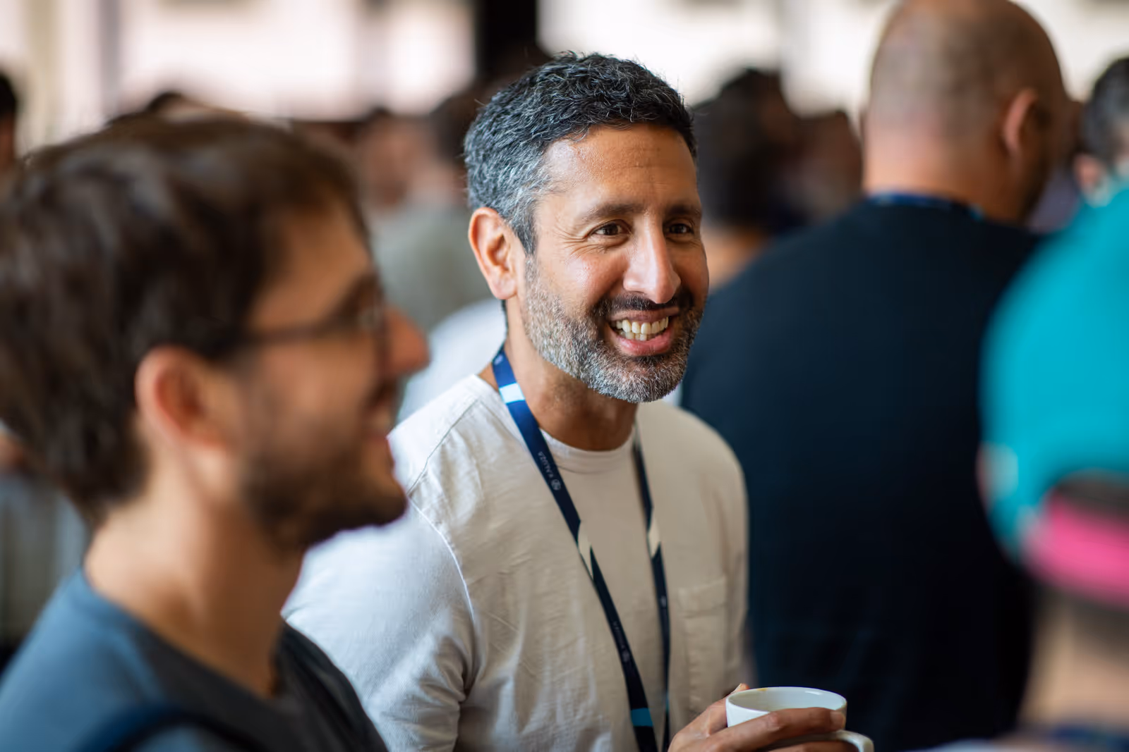 Two men smiling and talking at an indoor gathering, one holding a cup.