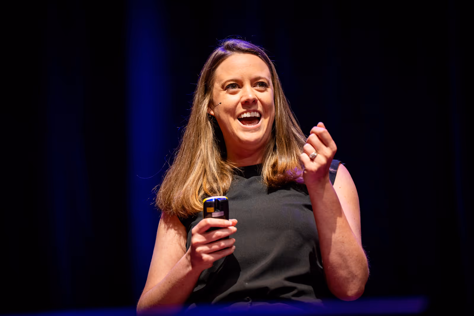 A woman smiles while speaking on stage, holding a small remote in her right hand.