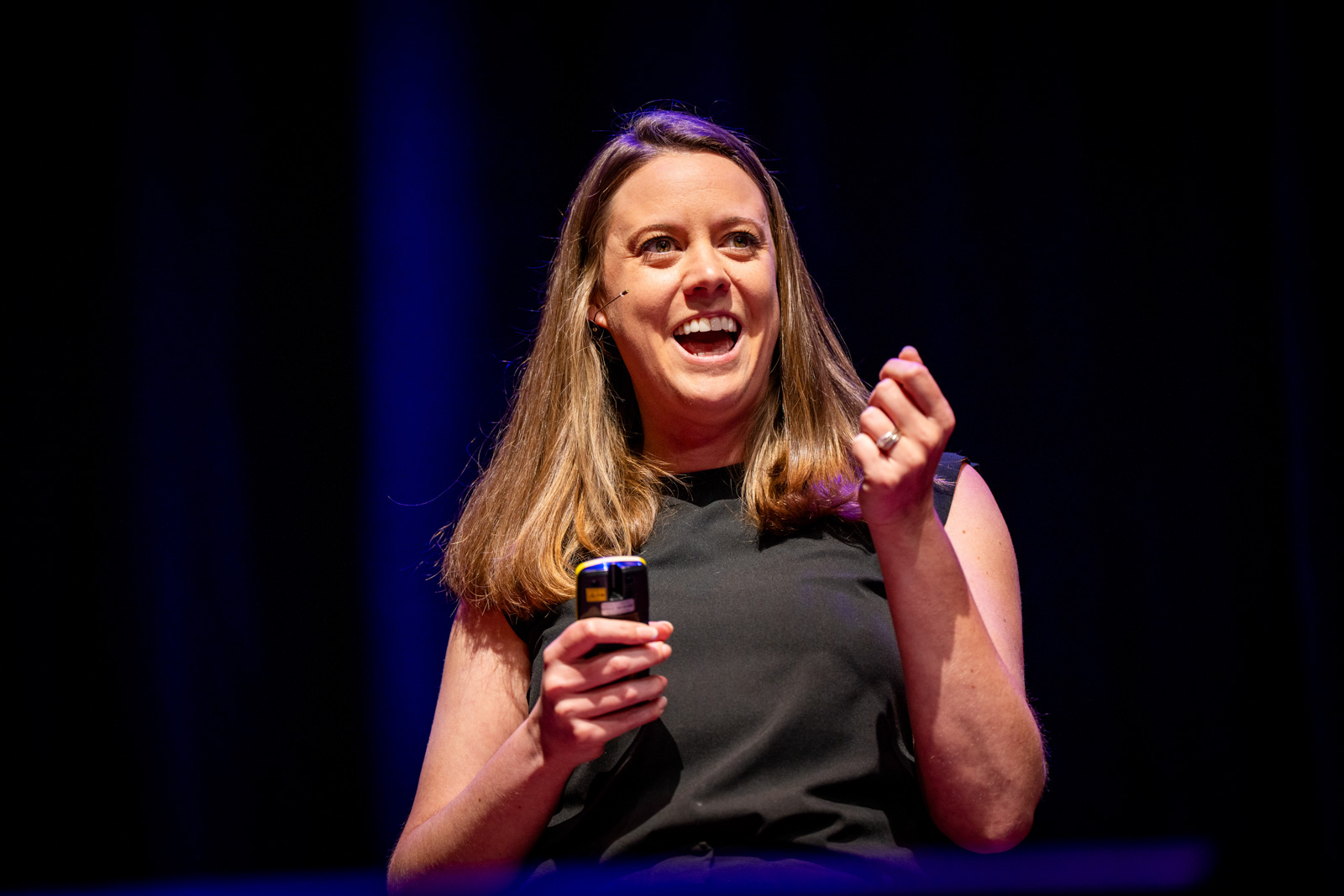 A woman smiles while speaking on stage, holding a small remote in her right hand.