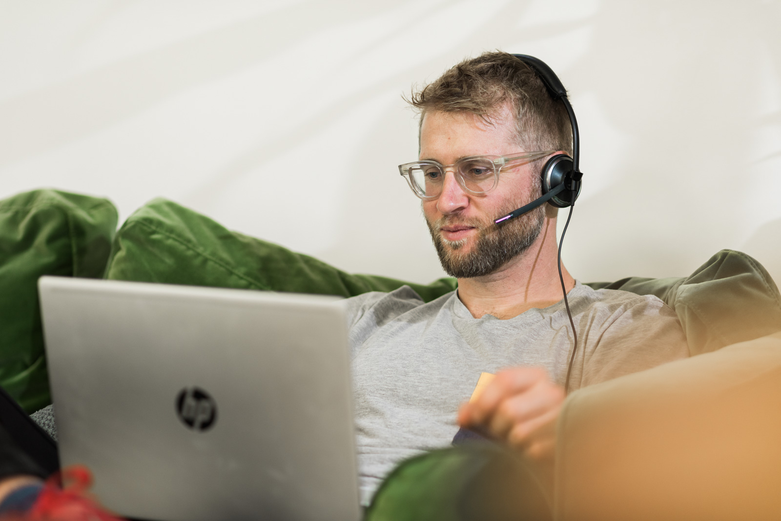 A man wearing a headset sits on a green couch, working on a laptop.
