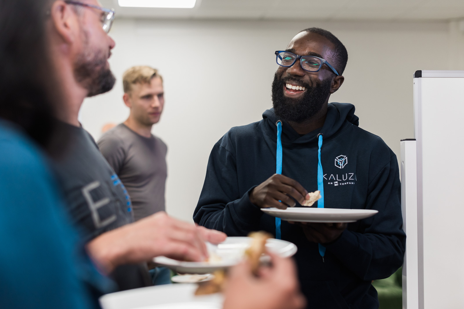 A man in a hoodie smiles while eating, standing with others at a casual gathering.