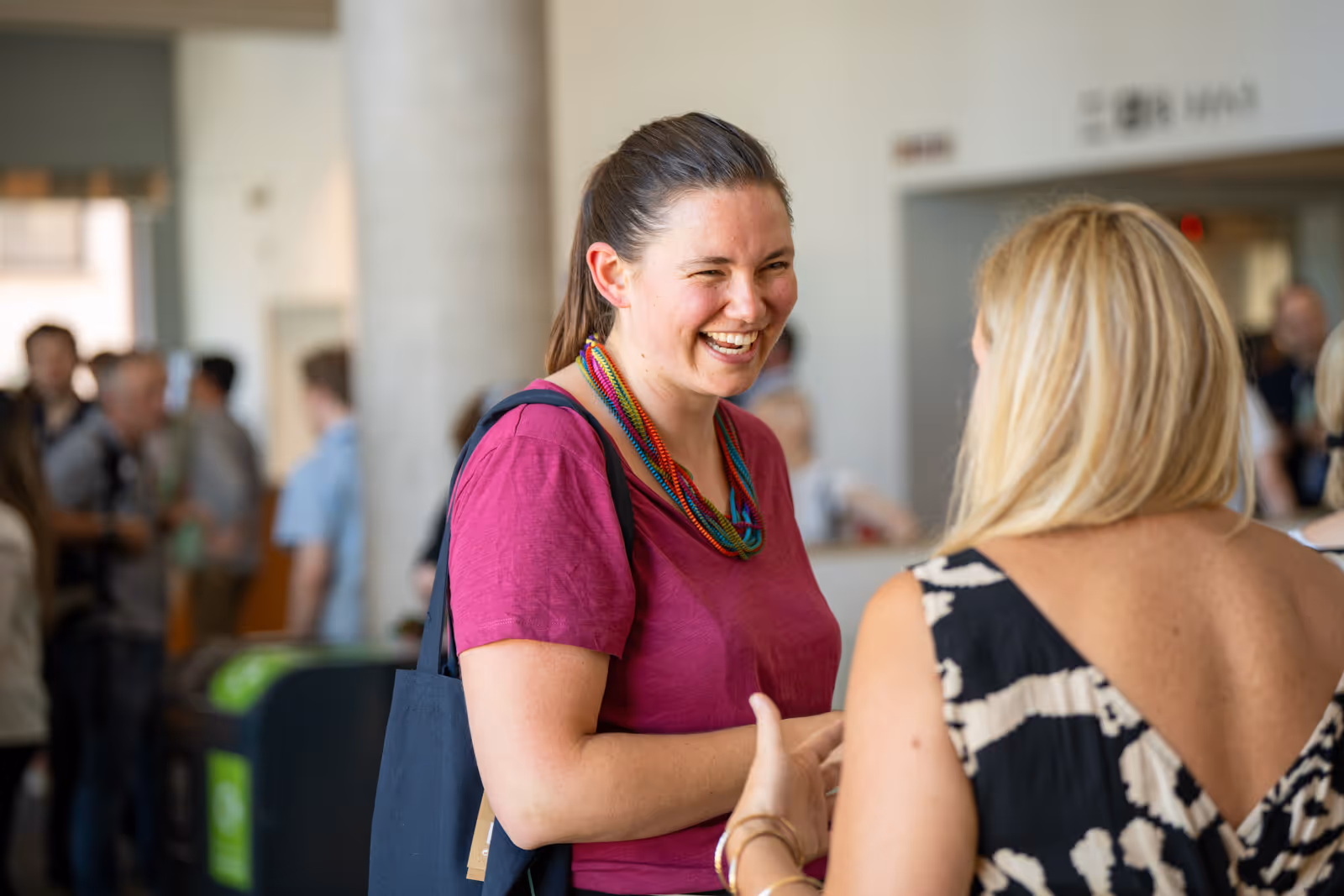 Two women are talking and smiling at each other in a busy indoor setting.
