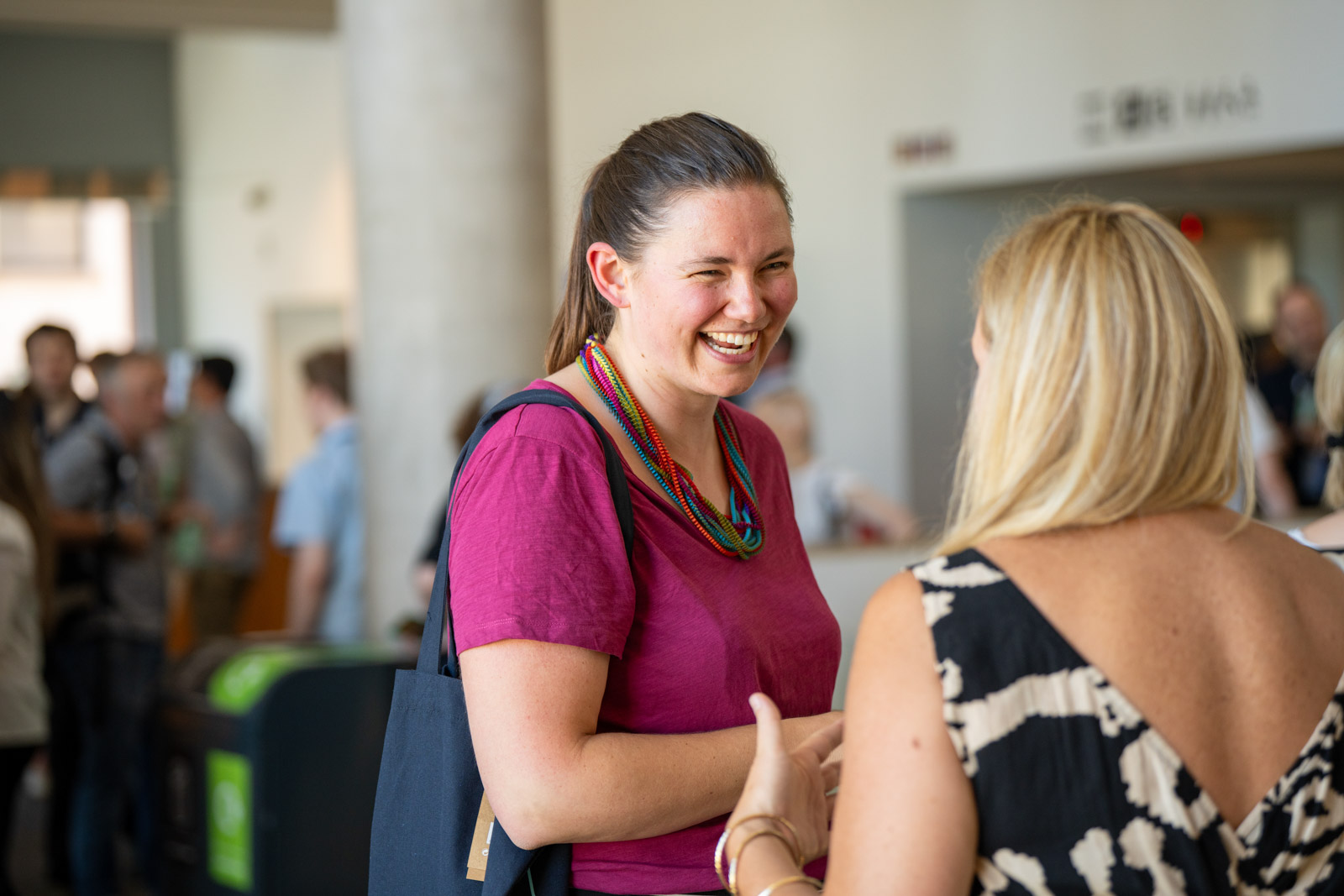 Two women are talking and smiling at each other in a busy indoor setting.