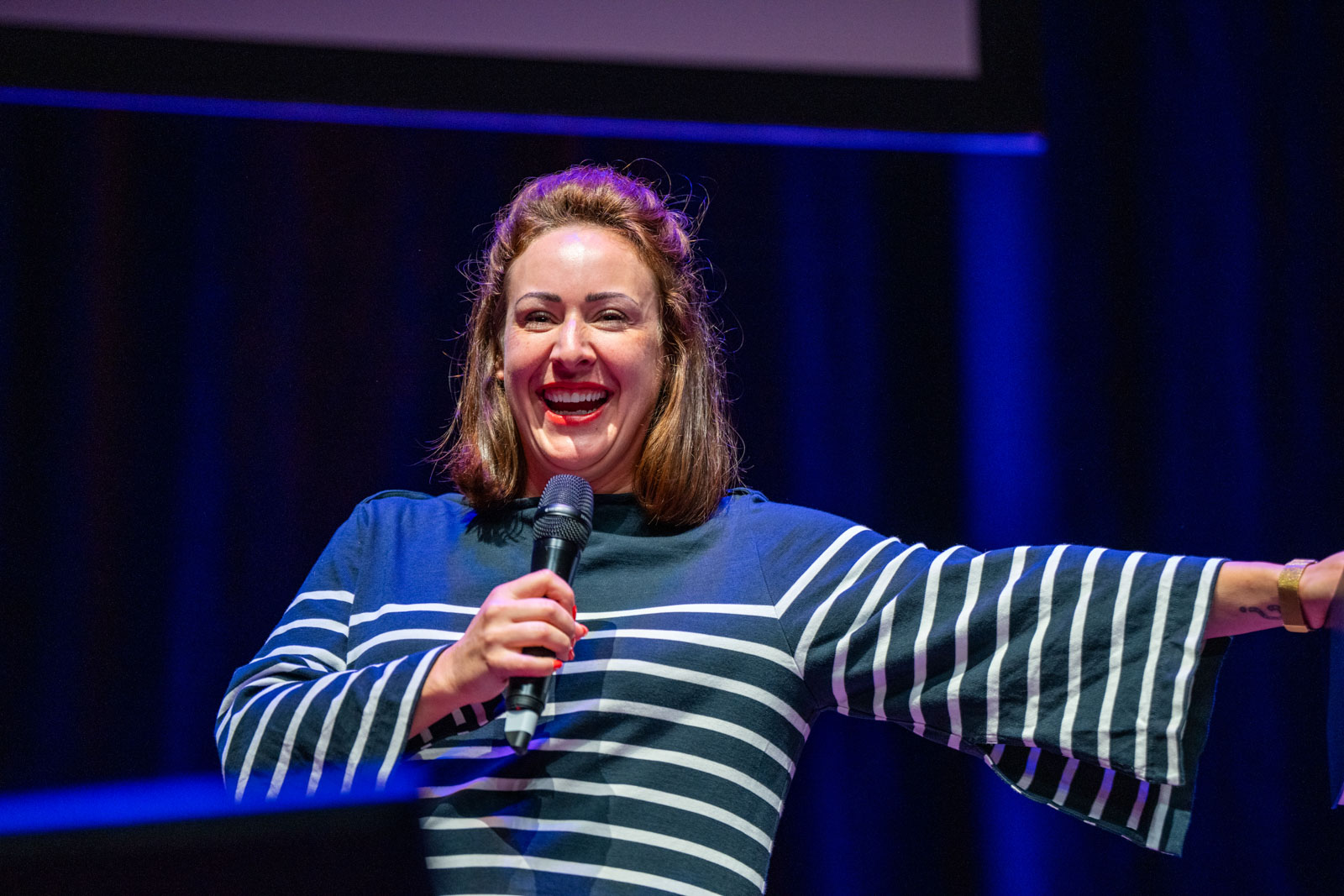 A person in a striped shirt is smiling and speaking into a microphone on stage.