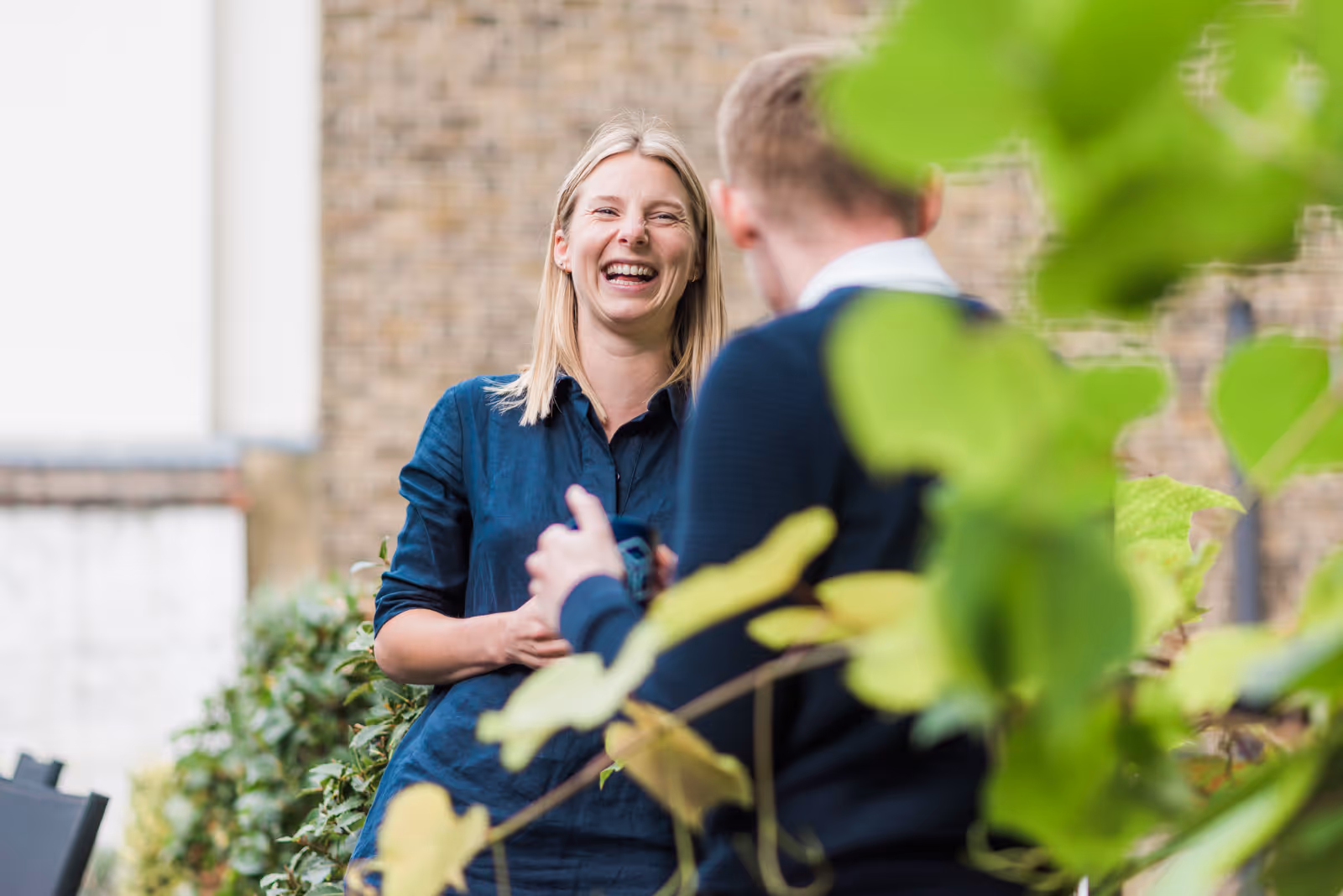 A woman in a blue shirt laughs while talking to a man outdoors.