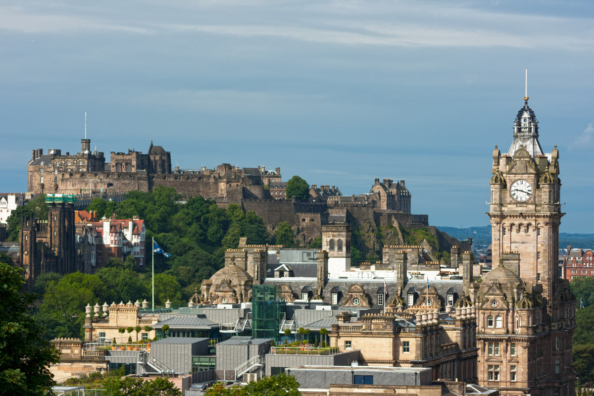 Castle and clock tower on a hill with green trees surrounding them under a blue sky.
