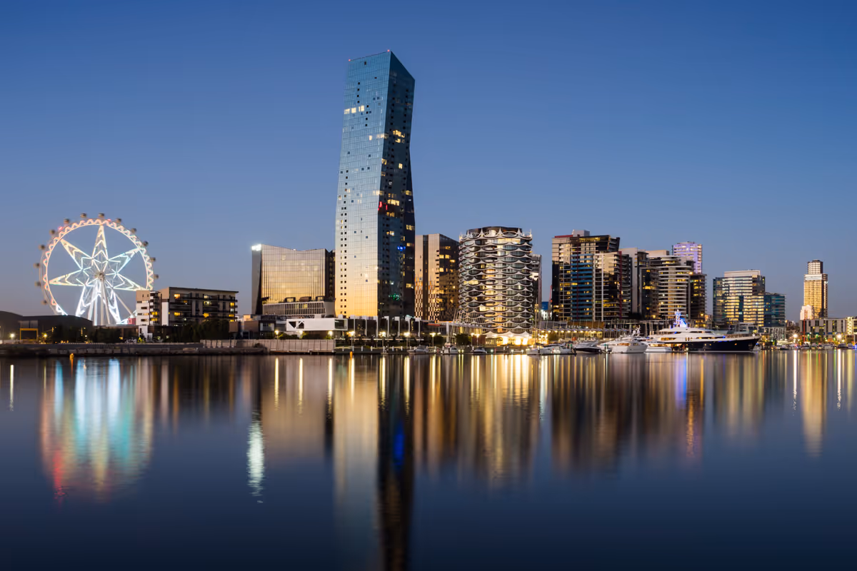 City skyline with illuminated Ferris wheel and tall buildings reflected in calm water at twilight.