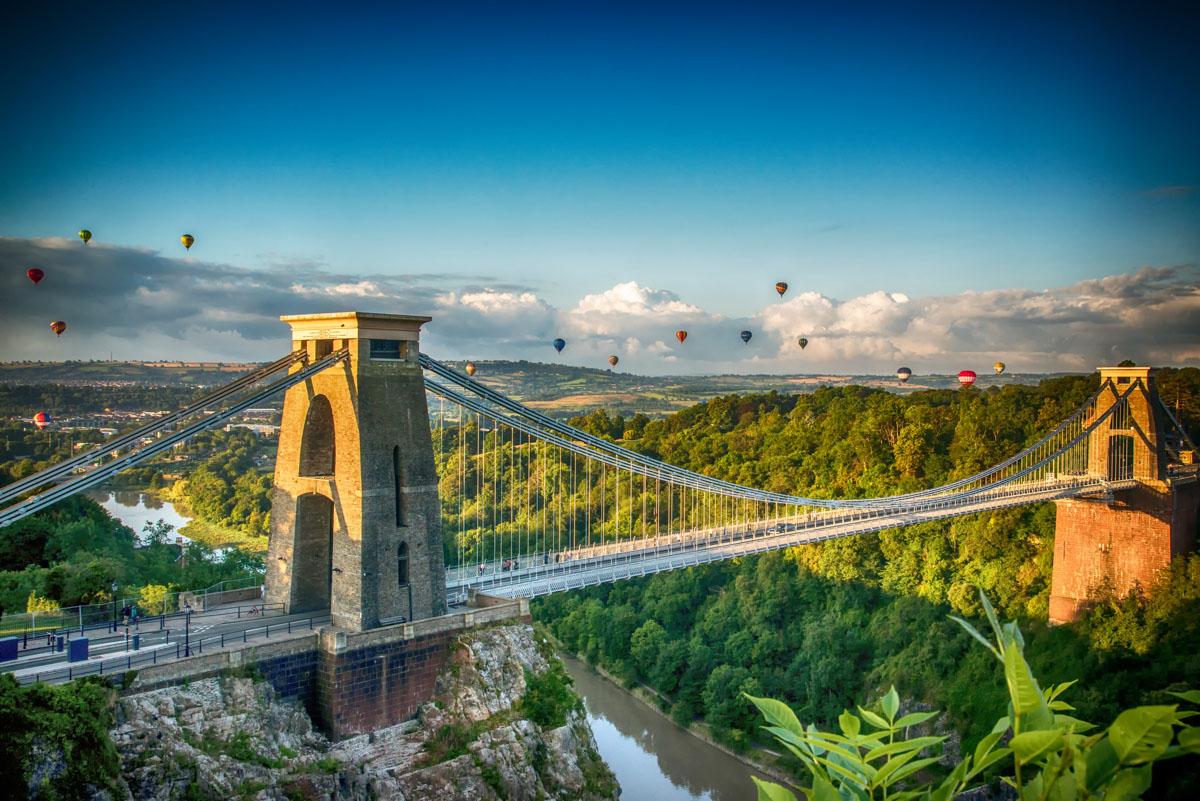 A suspension bridge spans over a river with several hot air balloons in the sky.