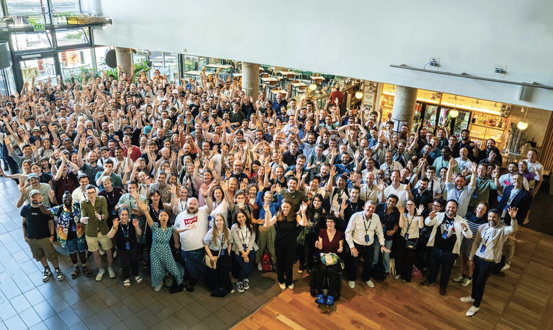 A large group of people stand together indoors, with many smiling and raising their hands.