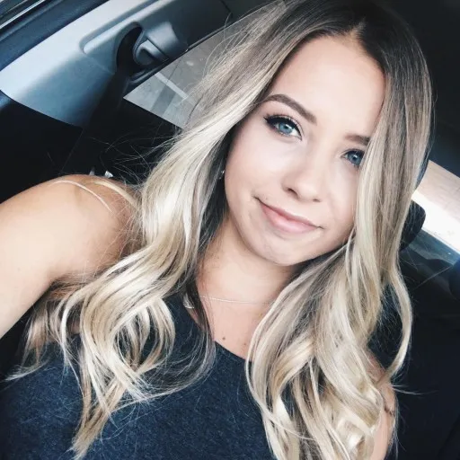 Young woman with long blonde wavy hair and blue eyes sitting in a car, smiling slightly at the camera.