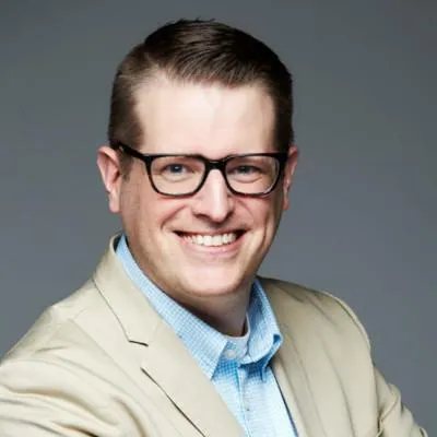 Smiling man with short brown hair, black glasses, wearing a beige blazer and blue checkered shirt against gray background.