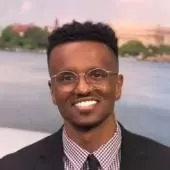 Smiling man with short curly hair and glasses, wearing a black suit and checkered dress shirt, standing outdoors with water and greenery in the background.