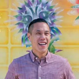 Smiling man wearing a light purple button-up shirt standing in front of a colorful mural.