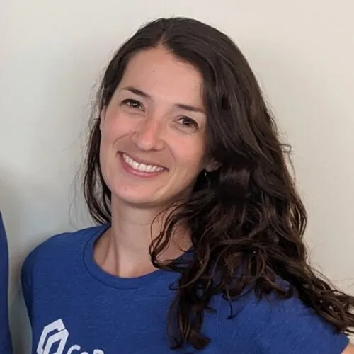 Woman with long dark curly hair smiling, wearing a blue shirt with white text on it.