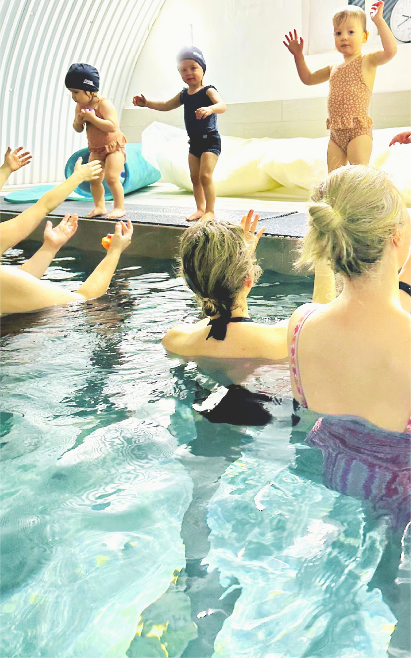 Three toddlers in swim caps stand at the pool edge while adults in swimsuits reach out from the water.