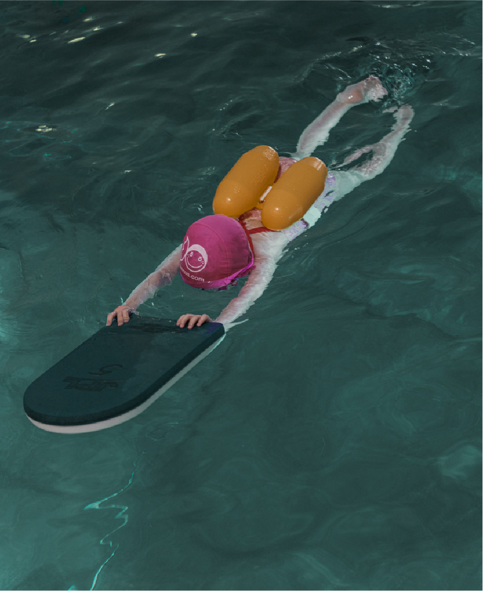 Child wearing a pink swim cap and orange floatation aids practicing swimming with a kickboard in a pool.